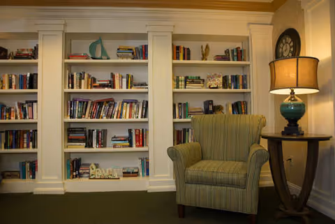 Cozy reading nook with a green striped armchair next to a wooden side table with a lamp. Behind the chair is a built-in white bookshelf filled with books and decorative items, and a large clock is mounted on the wall above the table.