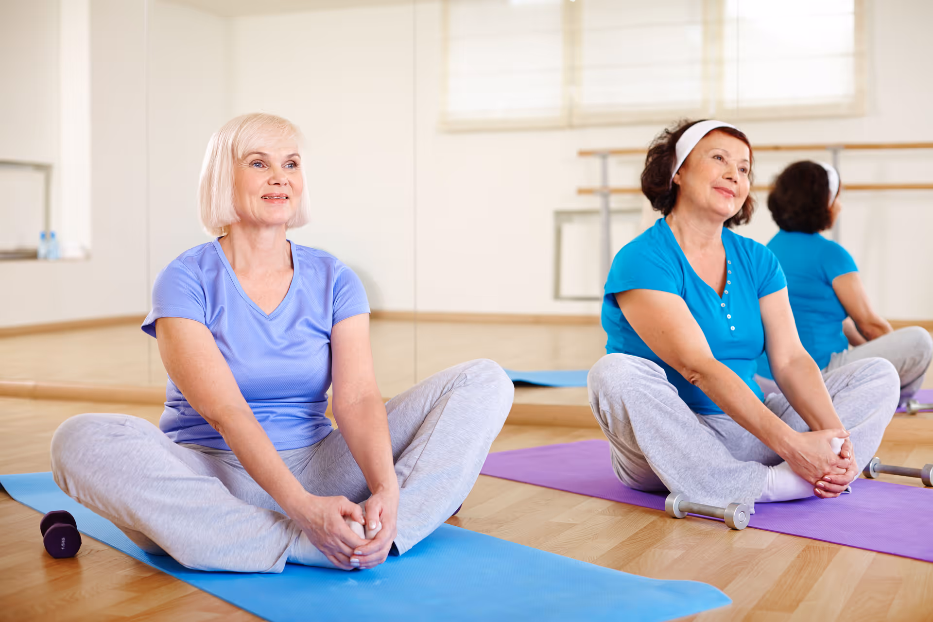 Two older women sitting on yoga mats in a fitness studio, stretching with their legs bent and hands holding their feet. They are dressed in comfortable workout clothes and appear relaxed and content. The room has wooden floors, a large mirror on the wall, and exercise equipment like dumbbells nearby.