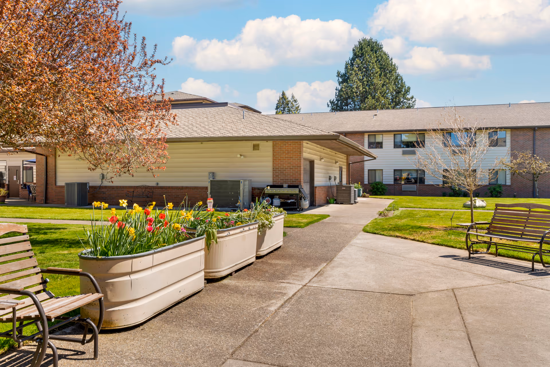 Outdoor courtyard area of Brookdale McMinnville Town Center featuring paved walkways, benches, flower planters with blooming flowers, a tree with red leaves, and a building with windows and a sloped roof under a partly cloudy sky.