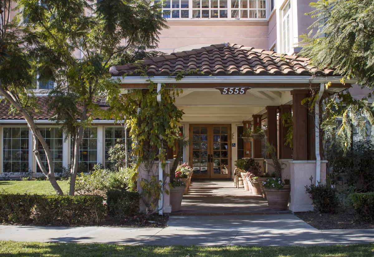 Entrance of a senior living facility with a covered walkway supported by wooden pillars and a tiled roof. The building number 5555 is displayed above the entrance. The area is surrounded by greenery, including trees, bushes, and potted plants lining the walkway leading to double glass doors.