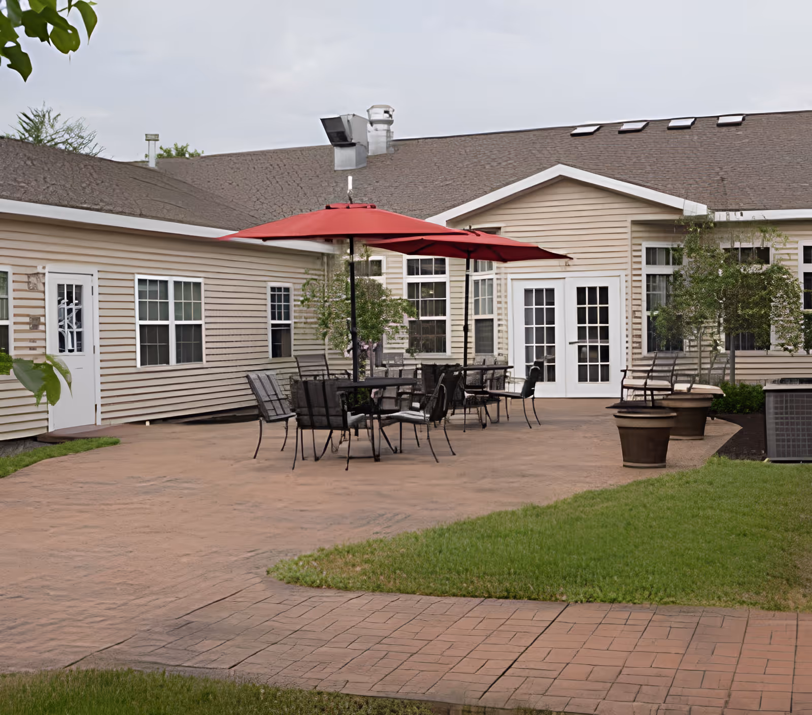 Outdoor patio area at a senior living facility with several black metal tables and chairs, two large red umbrellas providing shade, beige siding building with multiple windows and white doors in the background, and some potted plants and green grass surrounding the patio.