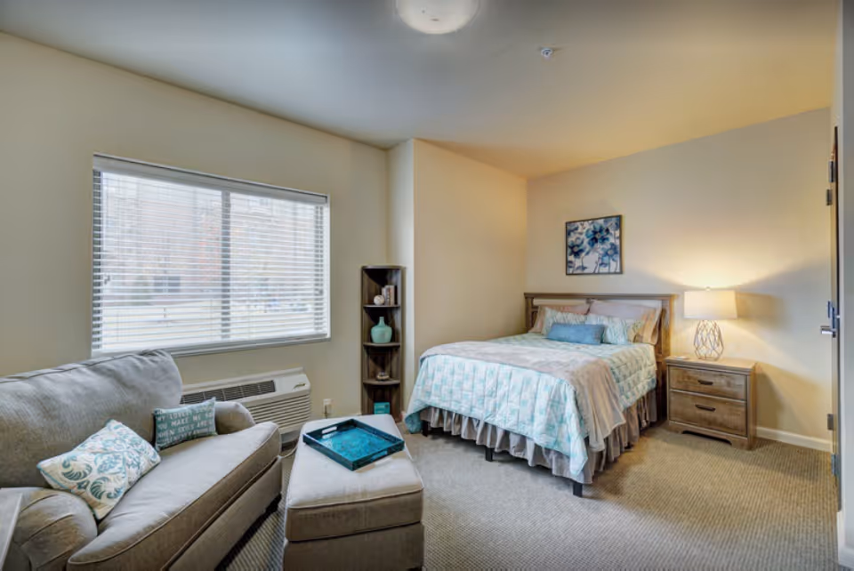 A cozy senior living bedroom with a bed covered in light blue and beige bedding, a wooden nightstand with a lamp, a small corner shelf with decorative items, a gray sofa with patterned pillows, an ottoman with a tray on top, and a large window with blinds letting in natural light.