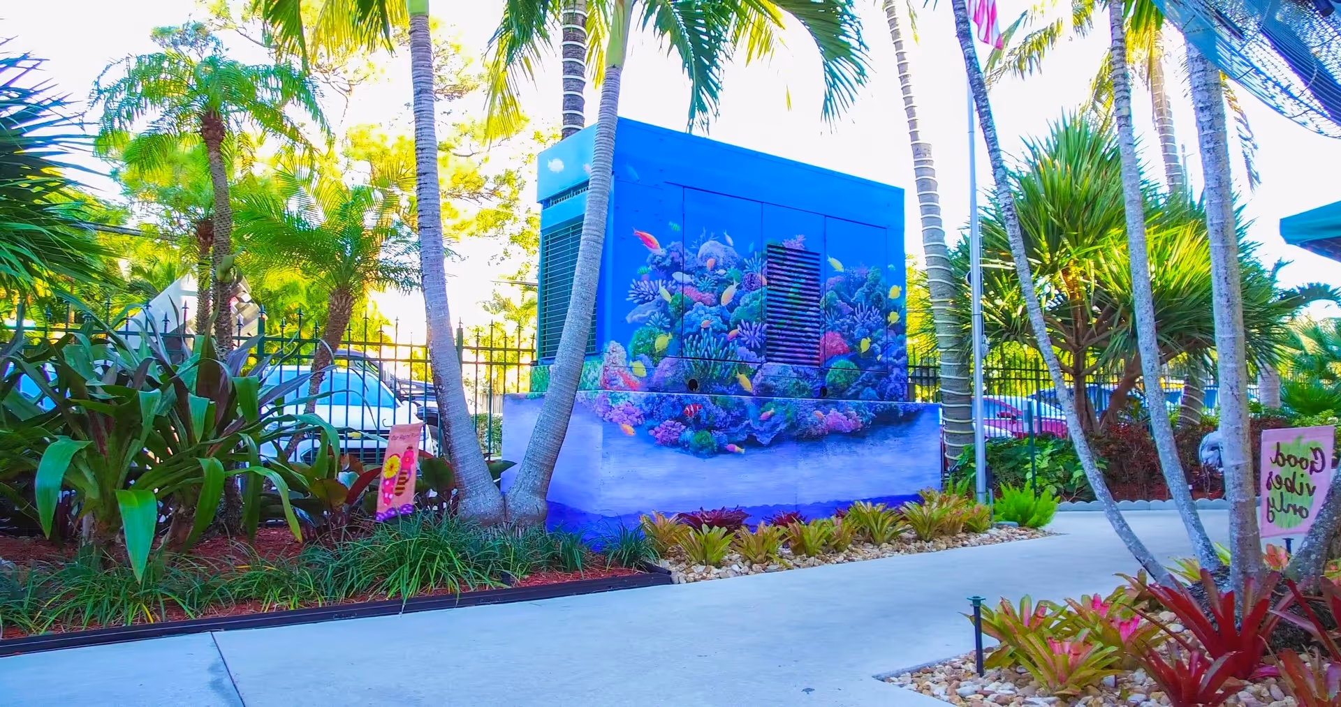 Tropical outdoor courtyard with palm trees and a brightly painted blue mural-covered utility box depicting a coral reef next to a paved walkway.