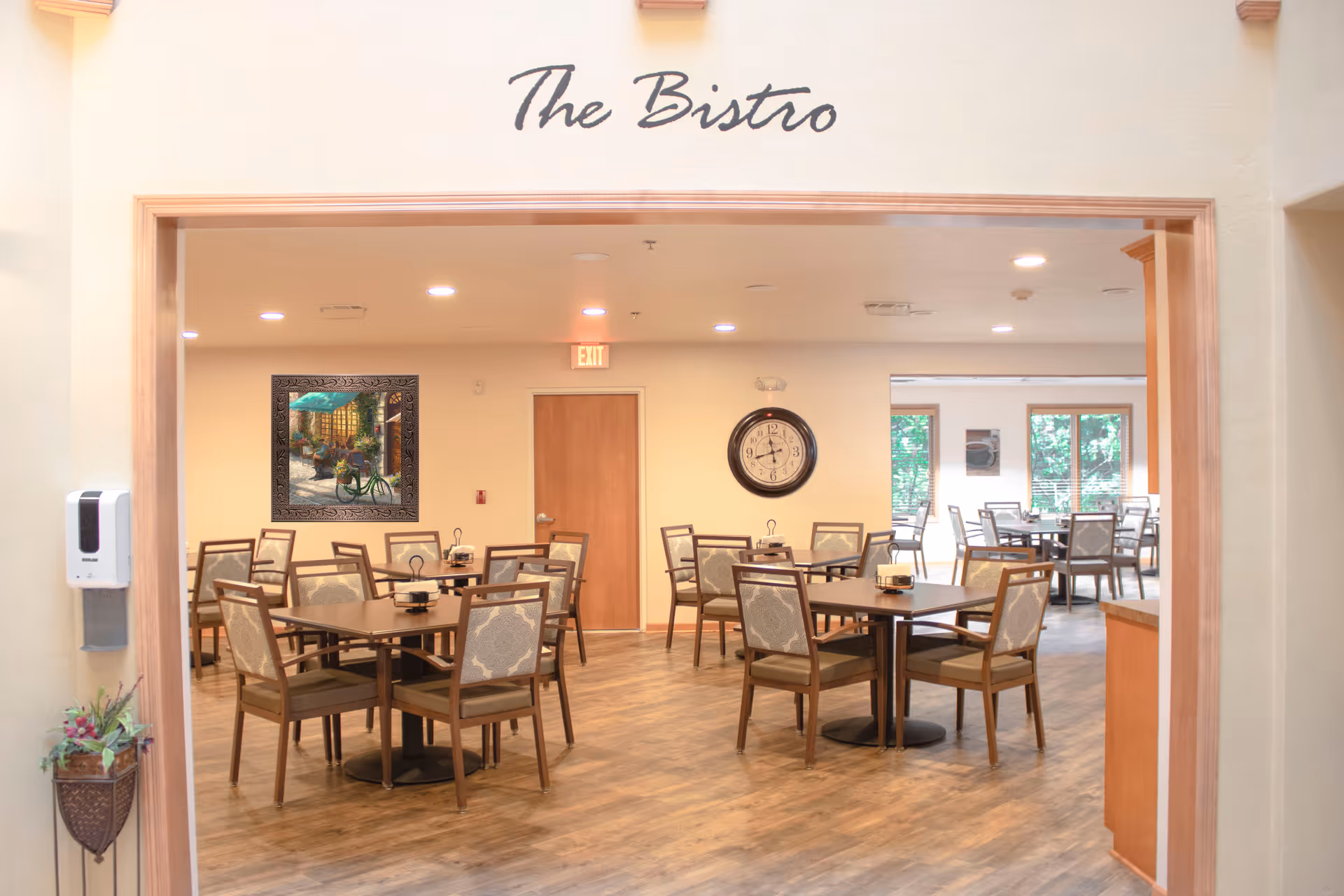 Interior view of a dining area named The Bistro with multiple tables and chairs arranged neatly. The room has wooden flooring, a wall clock, a painting, and large windows letting in natural light.