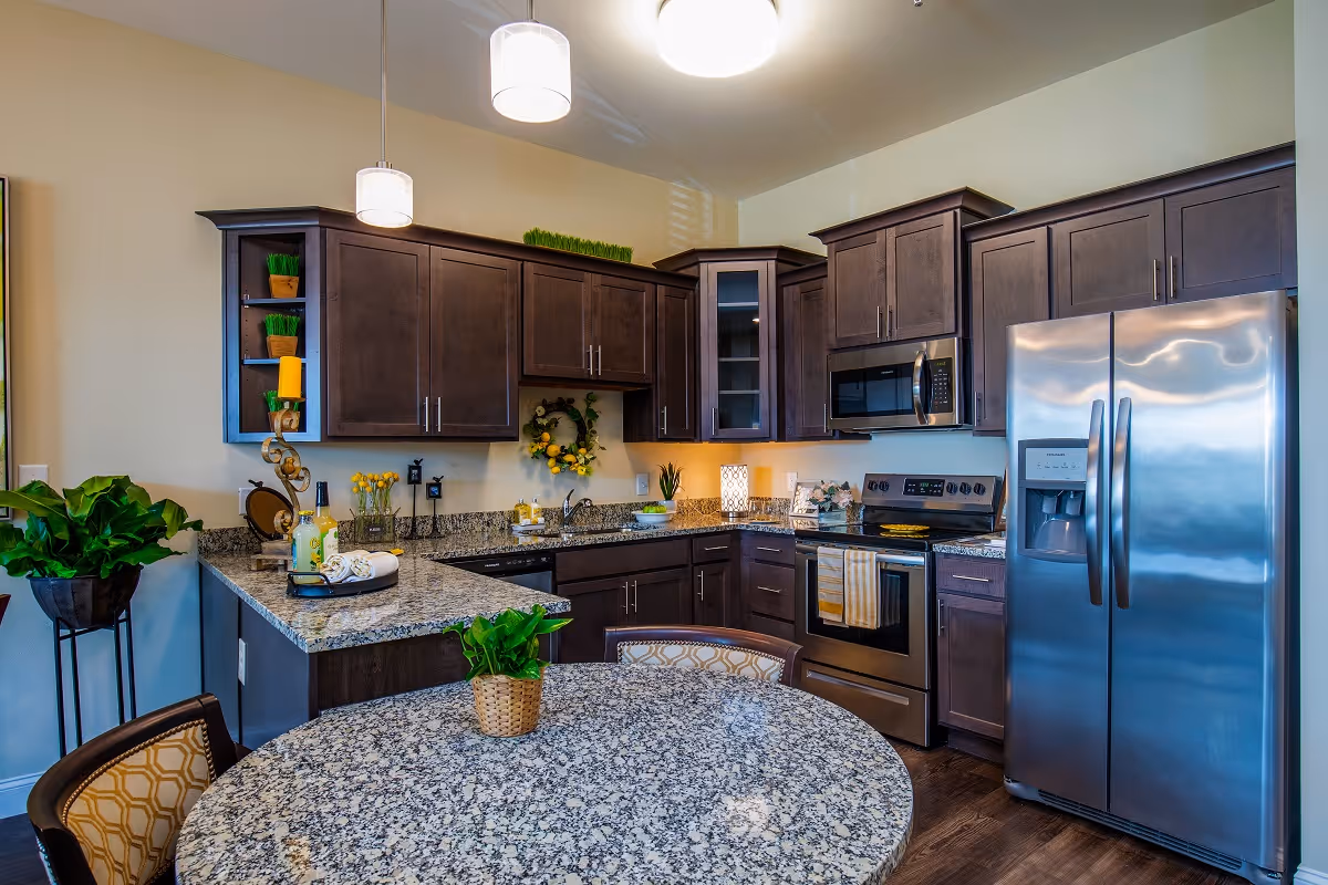 Modern kitchen with dark wood cabinets, granite countertops, stainless steel refrigerator and oven, a round granite table with chairs, and decorative plants and items on the counters and shelves.