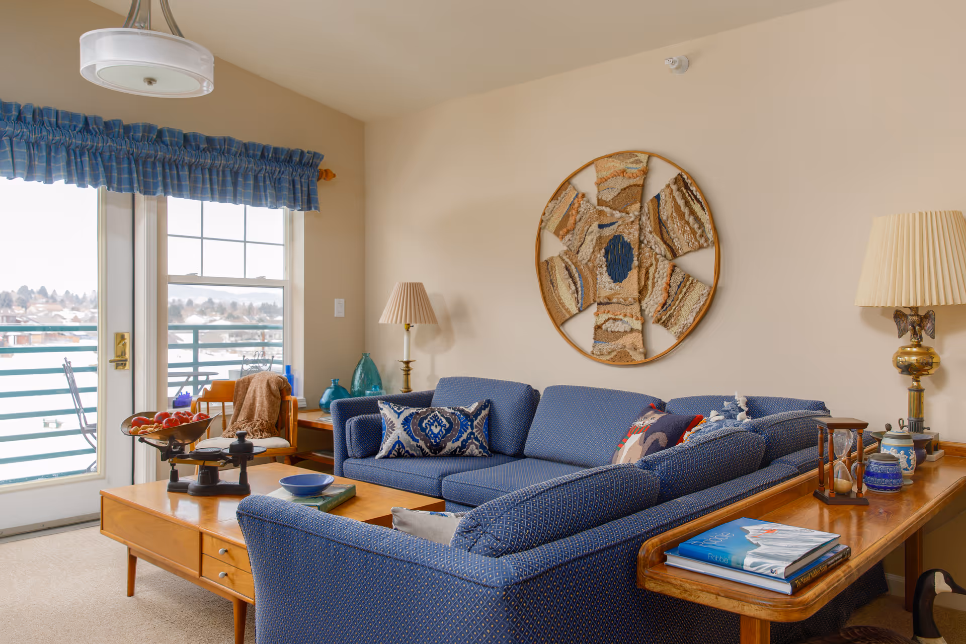 A cozy living room with a blue patterned sectional sofa adorned with decorative pillows. A wooden coffee table in front of the sofa holds a bowl of apples, a book, and a decorative scale. Behind the sofa is a wooden side table with books, an hourglass, and a lamp with a pleated shade. On the wall above the sofa is a large circular textile art piece. To the left, there is a window and a glass door with blue plaid valances, leading to an outdoor balcony with a railing and a view of trees and buildings in the distance.
