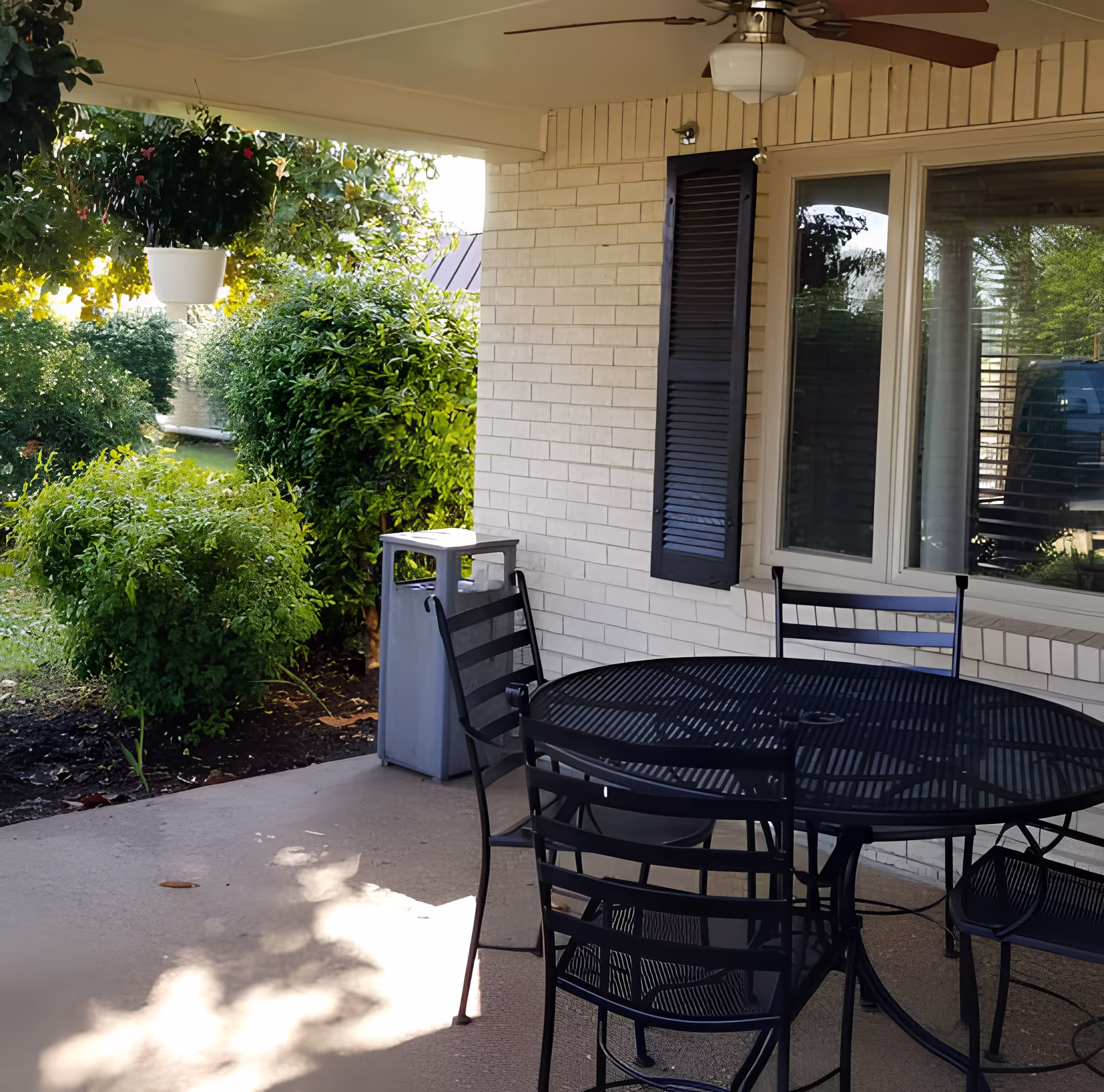 Outdoor covered patio area with a round black metal table and four matching chairs. There is a ceiling fan with a light above the table. The patio is adjacent to a light-colored brick wall with a window and black shutters. Lush green bushes and plants surround the patio area.