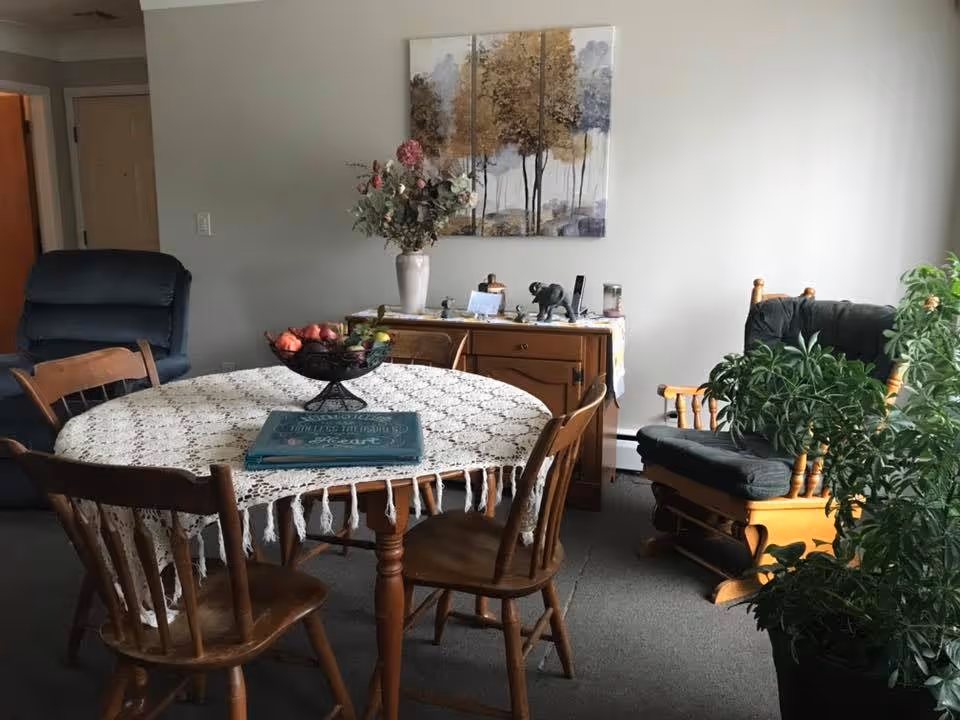 A cozy interior room featuring a round wooden dining table covered with a white lace tablecloth, surrounded by four wooden chairs. On the table is a decorative bowl with fruit and a book. In the background, there is a wooden sideboard with a vase of flowers and various small decorative items. To the right, there is a cushioned wooden rocking chair next to a large green potted plant. A three-panel painting of trees hangs on the wall above the sideboard.