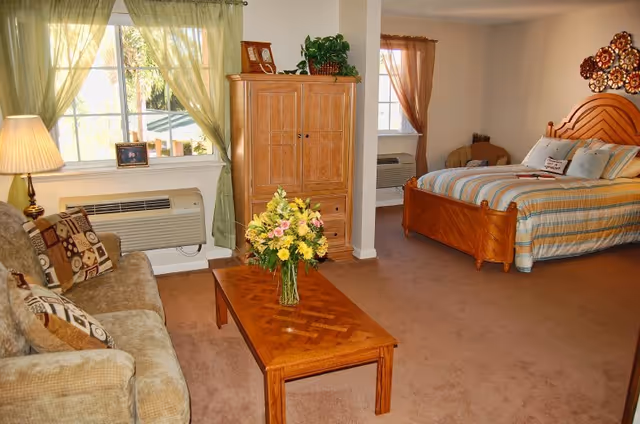 A cozy senior living facility room featuring a sitting area with a beige couch adorned with patterned pillows, a wooden coffee table with a vase of fresh flowers, a wooden armoire, and a bed with striped bedding. The room has windows with light green and brown curtains, a lamp on a side table, and carpeted flooring.