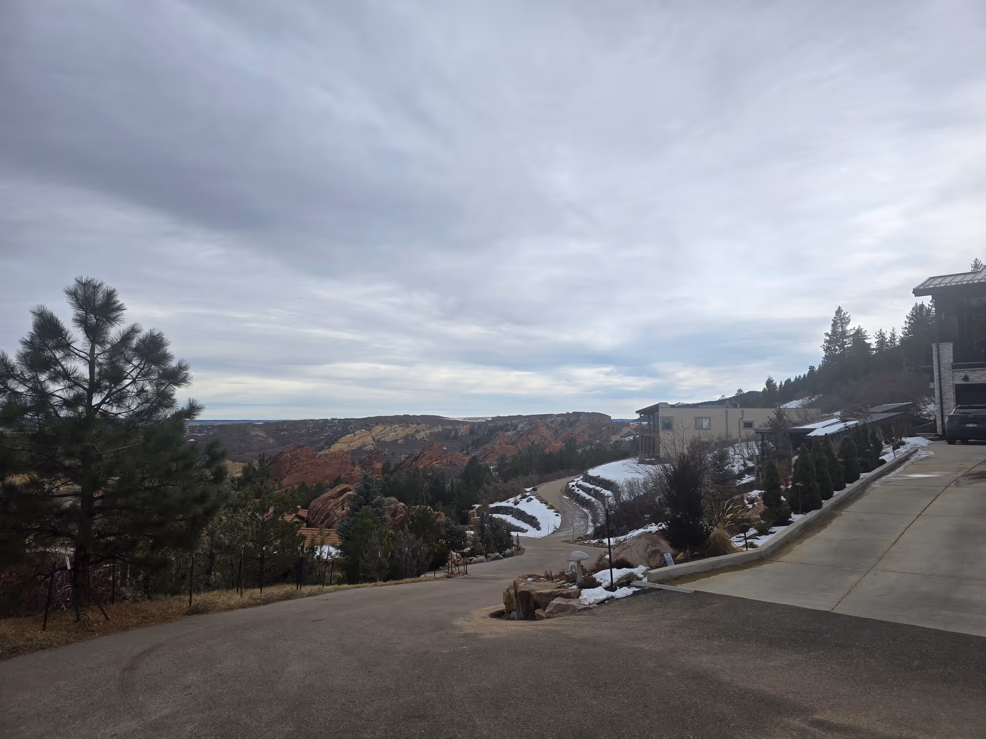 A winding paved road leading through a mountainous area with scattered snow patches. There are trees and shrubs along the road, with rocky hills and red rock formations visible in the background under a cloudy sky. A driveway and part of a building are visible on the right side of the image.