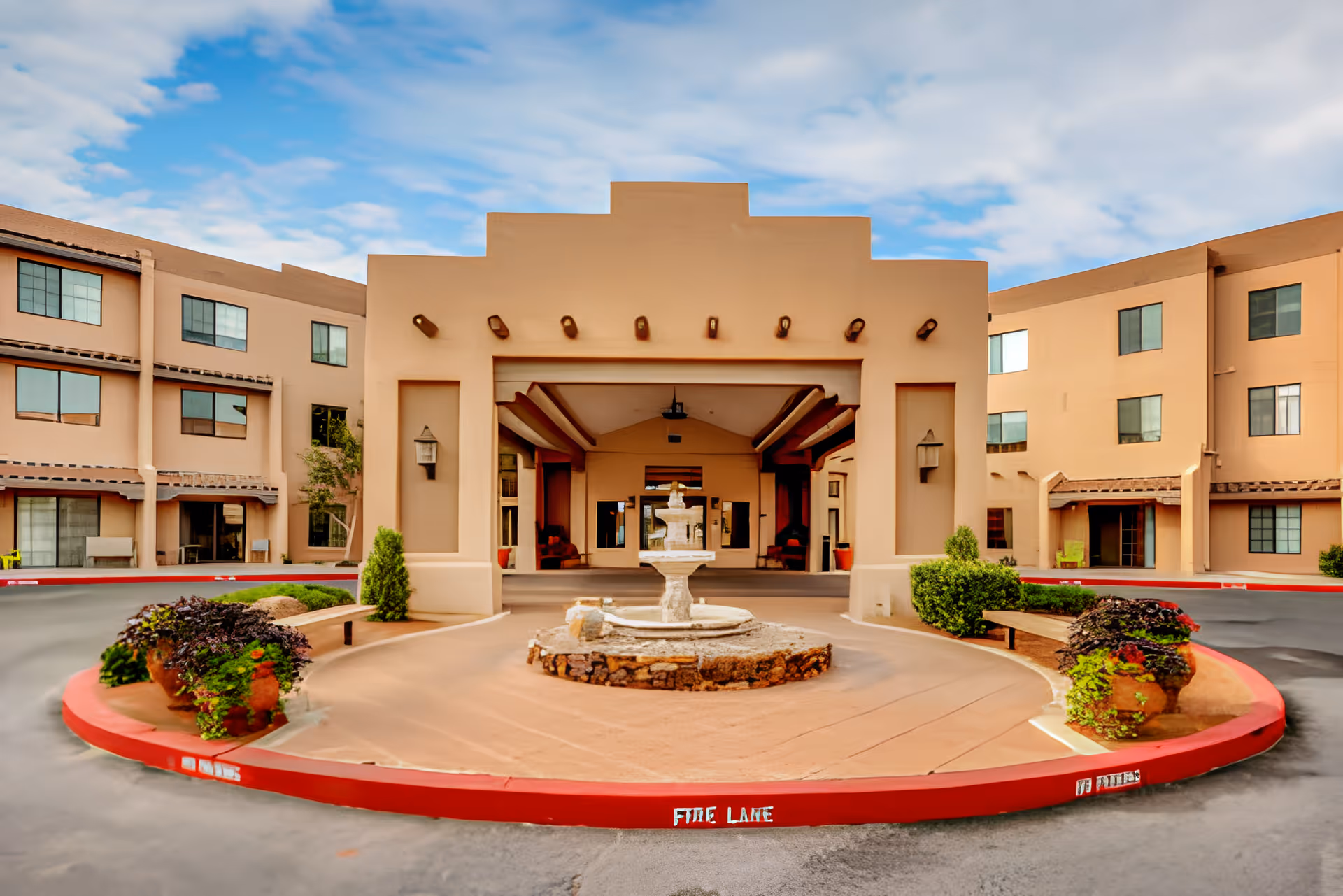 Front entrance of a senior living facility with a beige stucco exterior, a covered drop-off area, a central stone fountain, and landscaped planters with greenery. The building has multiple windows and a clear blue sky with some clouds in the background.