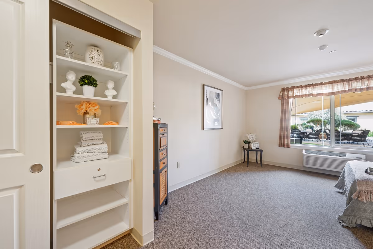 A bright and tidy room with beige walls and carpeted floor. On the left, there is a built-in shelving unit with decorative items including small busts, a potted plant, folded towels, and a flower arrangement. In the background, a framed picture hangs on the wall above a small table with a flower vase and a clock. A large window with plaid curtains lets in natural light and shows an outdoor patio area with tables and chairs. A bed with a textured blanket is partially visible on the right side.