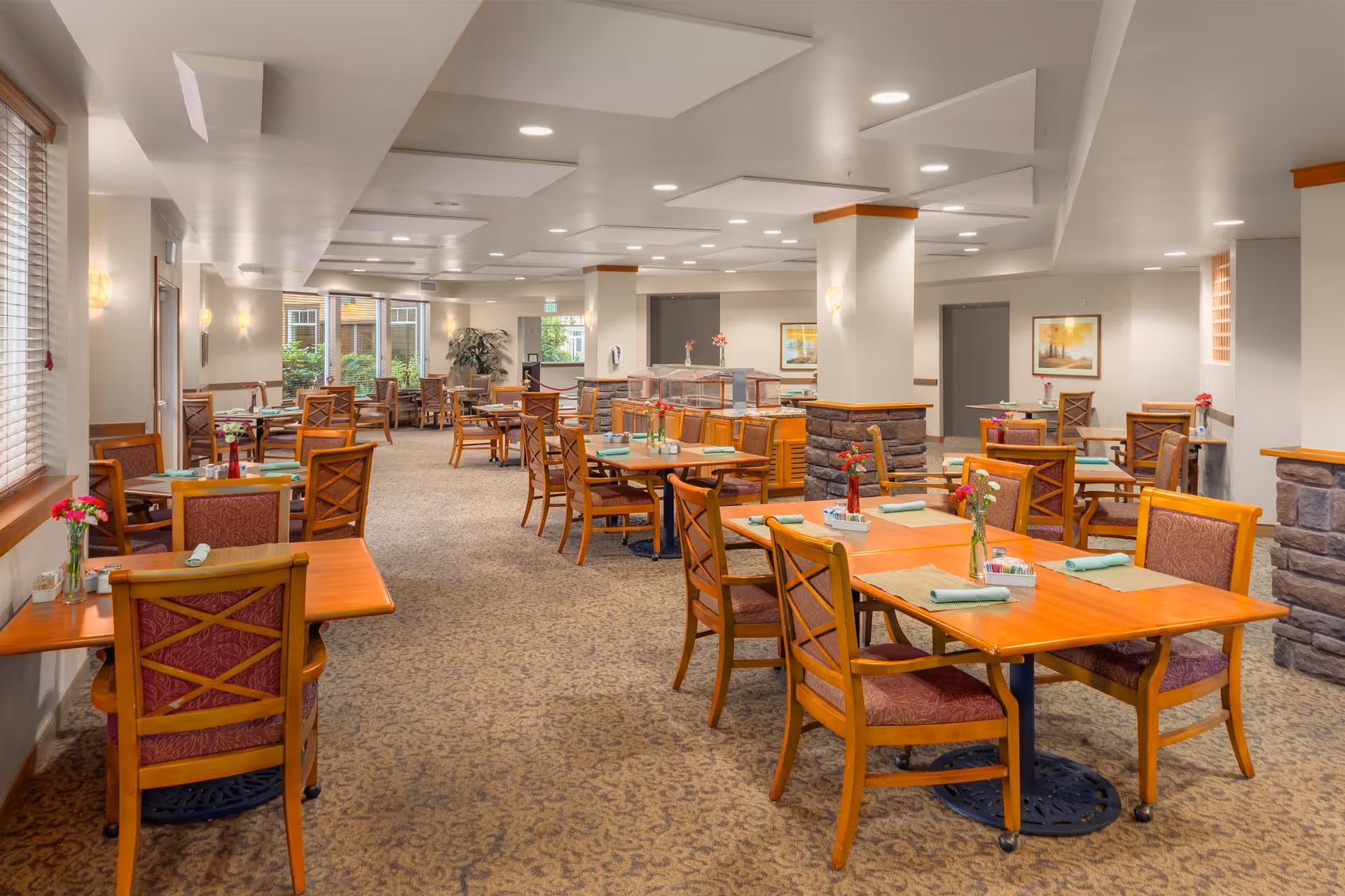 A spacious dining room with multiple wooden tables and chairs arranged neatly. Each table has placemats, rolled napkins, and small vases with flowers. The room has carpeted floors, light-colored walls, and ceiling lights. Large windows on the left side allow natural light to enter, and there are decorative stone pillars in the room.