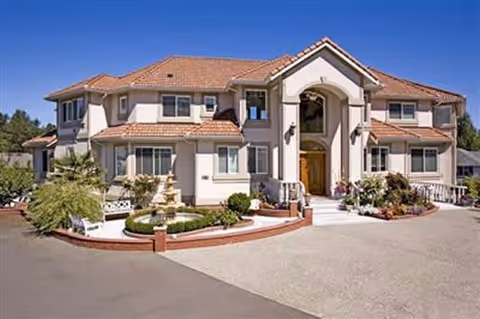 Large two-story residential building with a tiled roof, multiple windows, and a central entrance with an archway. The front yard features a circular garden with a fountain, shrubs, and flowers. The driveway and surrounding area are paved.
