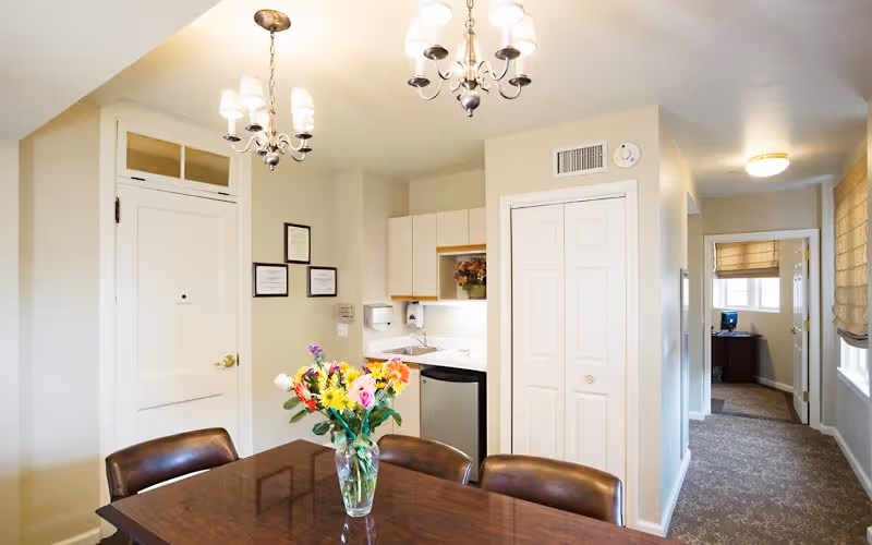 A bright interior space featuring a dining table with a vase of colorful flowers, surrounded by four brown leather chairs. In the background, there is a small kitchenette with white cabinets, a sink, and a mini refrigerator. The room has beige walls, two chandeliers hanging from the ceiling, and a hallway leading to another room with a desk and computer visible.