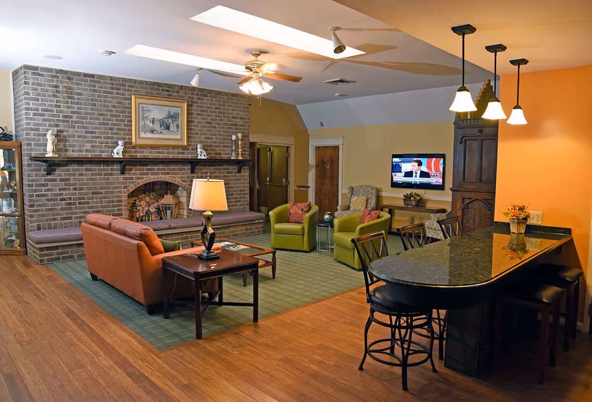 A cozy living room area with a brick fireplace, a ceiling fan, and a mounted TV. The room features a brown leather sofa, two green armchairs with red pillows, a wooden coffee table, and a side table with a lamp. There is a granite countertop bar with stools on the right side and warm yellow and orange walls.