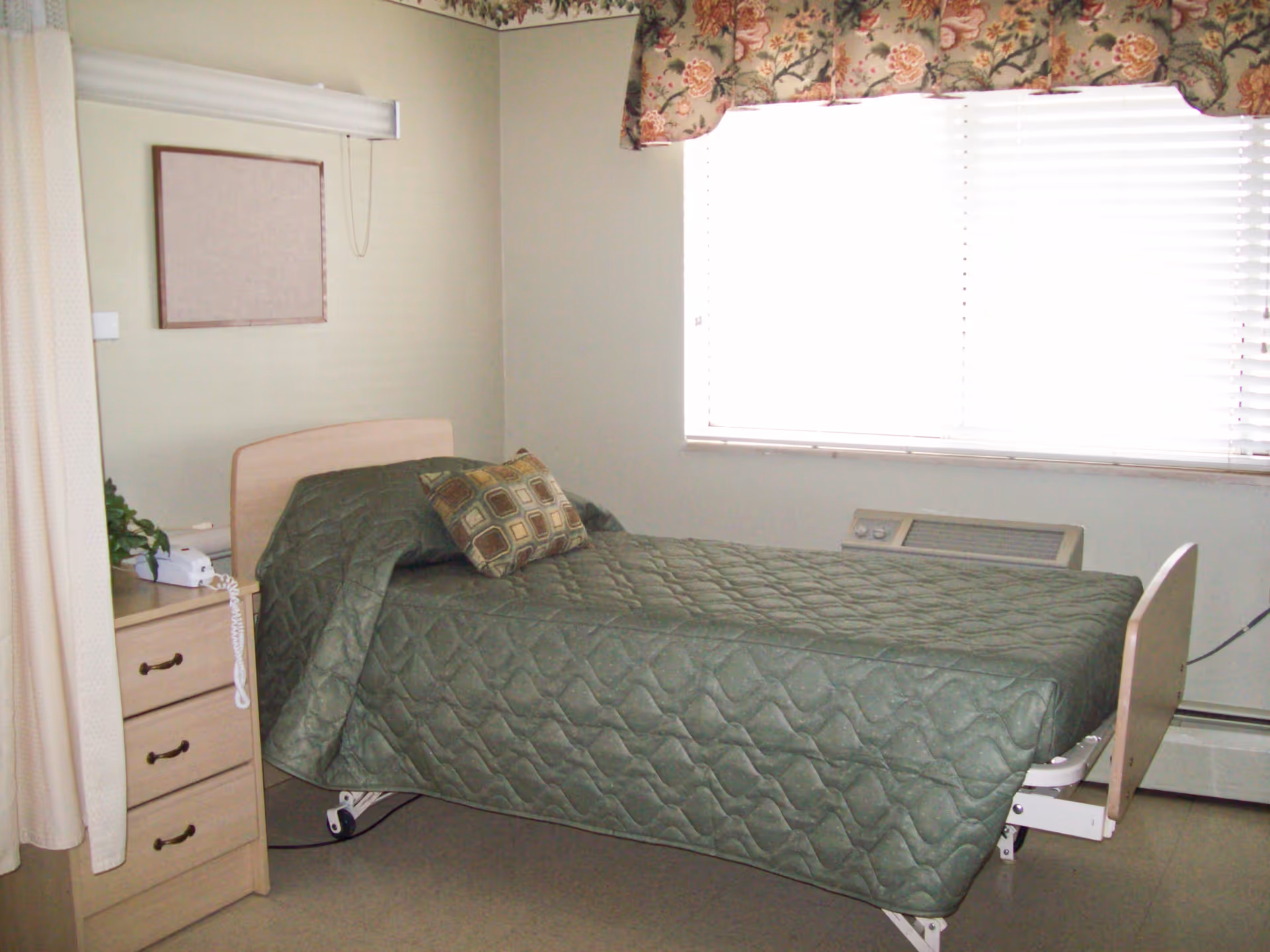A simple bedroom in a care facility with a single bed covered in a green quilted bedspread and a patterned pillow. Next to the bed is a wooden nightstand with three drawers, a white telephone, and a small plant. A window with white blinds and floral valance lets in natural light. The walls are light-colored and there is a framed corkboard above the nightstand.