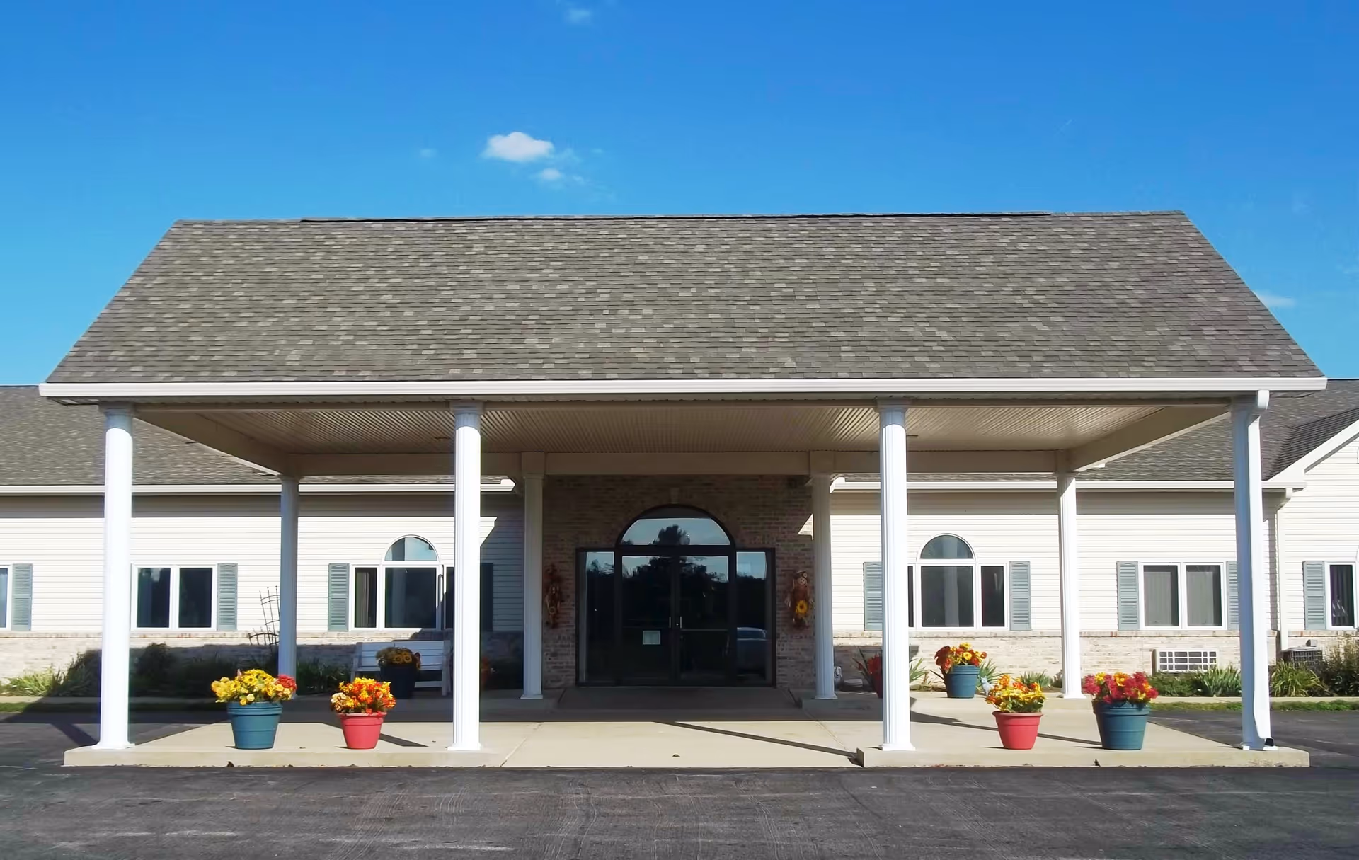 Front entrance of Eastland Retirement Community featuring a covered driveway supported by white columns, with potted flowers placed along the walkway and a clear blue sky overhead.