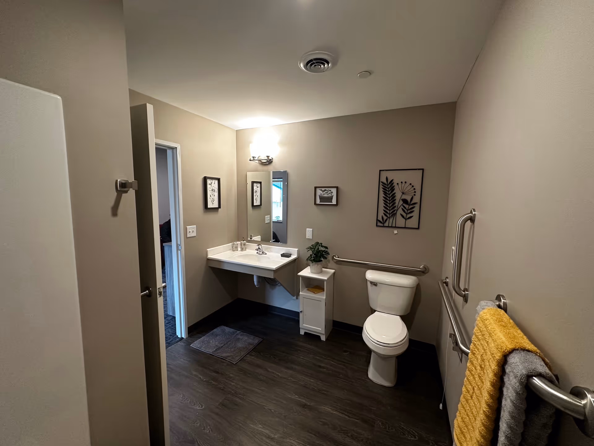 A clean, accessible bathroom with beige walls and dark wood flooring. The room features a white toilet with grab bars on both sides, a white sink with a mirror above it, a small white cabinet with a potted plant, and framed botanical artwork on the walls. There are two towels, one yellow and one gray, hanging on a grab bar on the right wall.