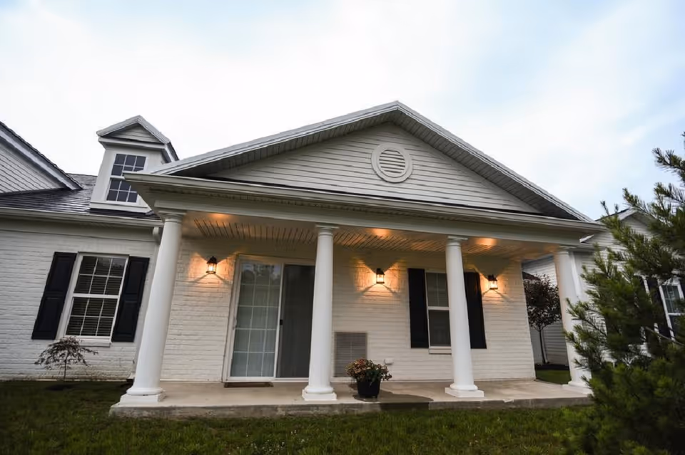 Exterior view of a single-story white brick building with a covered porch supported by four white columns. The porch has three wall-mounted lantern-style lights illuminating the area. There are two windows with black shutters and a sliding glass door. The building is surrounded by grass and some small trees and shrubs.