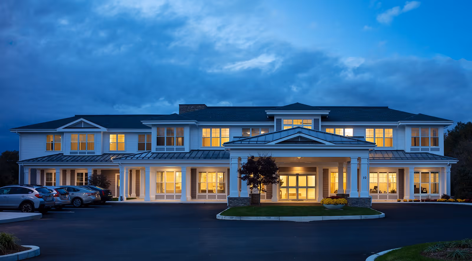 Exterior view of Wingate Residences at Haverhill building at dusk with lights on inside, a covered entrance with columns, several parked cars in front, and a cloudy sky overhead.