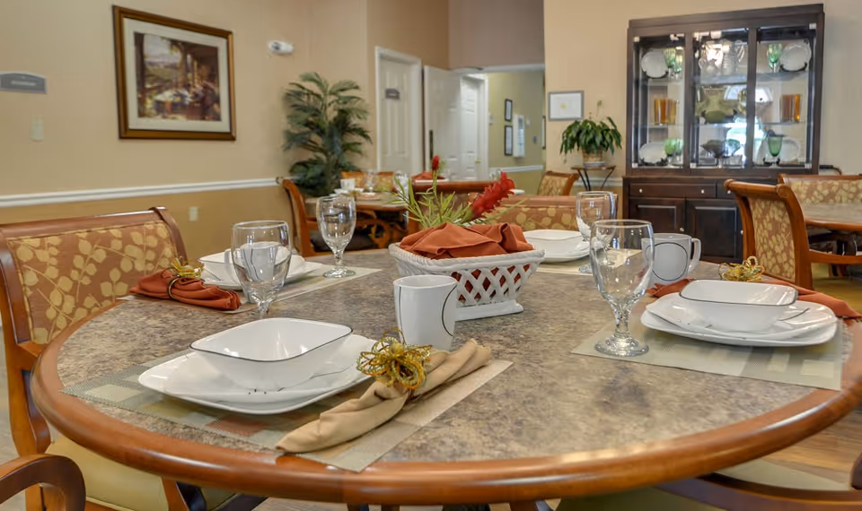 A dining room table set for four with plates, bowls, glasses, mugs, and cloth napkins with decorative napkin rings. The table has a centerpiece with a basket and red flowers. In the background, there are more tables and chairs, a plant, a framed picture on the wall, and a cabinet displaying glassware and dishes.
