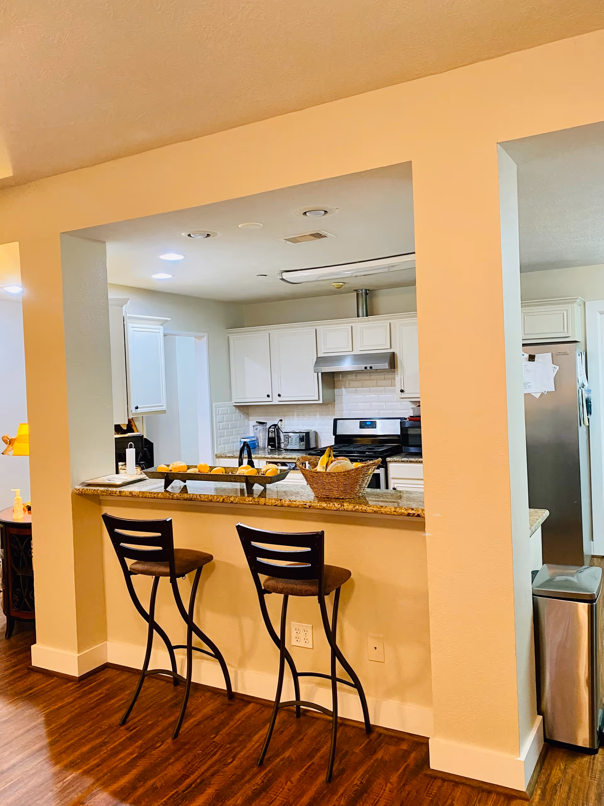 Interior view of a kitchen with white cabinets, a stainless steel stove and refrigerator, and a granite countertop bar with two high chairs. The floor is wooden, and there is a basket of fruit on the counter.