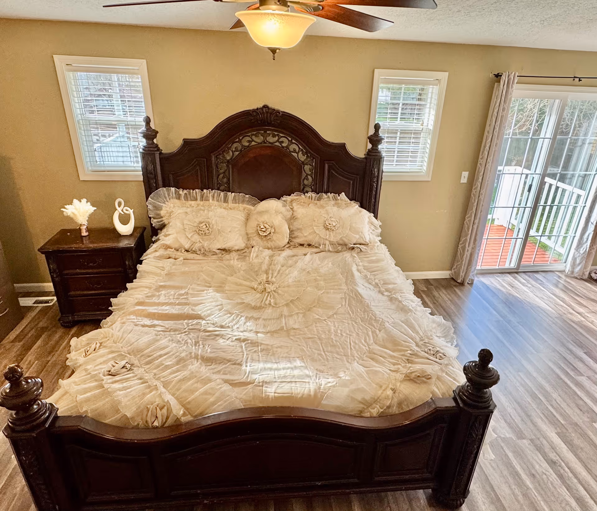 A bedroom with a large dark wooden bed frame featuring intricate carvings. The bed is made with a cream-colored, ruffled bedspread and matching decorative pillows. There are two small windows with white blinds on the wall behind the bed, a wooden nightstand with decorative items on the left side, and a sliding glass door with curtains on the right side leading to an outdoor area. The room has wood flooring and a ceiling fan with a light fixture.