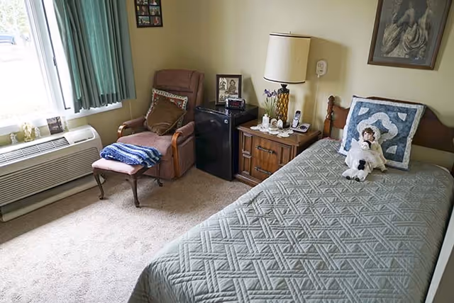 Small bedroom with a single bed, quilted bedspread and doll, a recliner chair by a curtained window, nightstand with lamp and a mini-fridge.