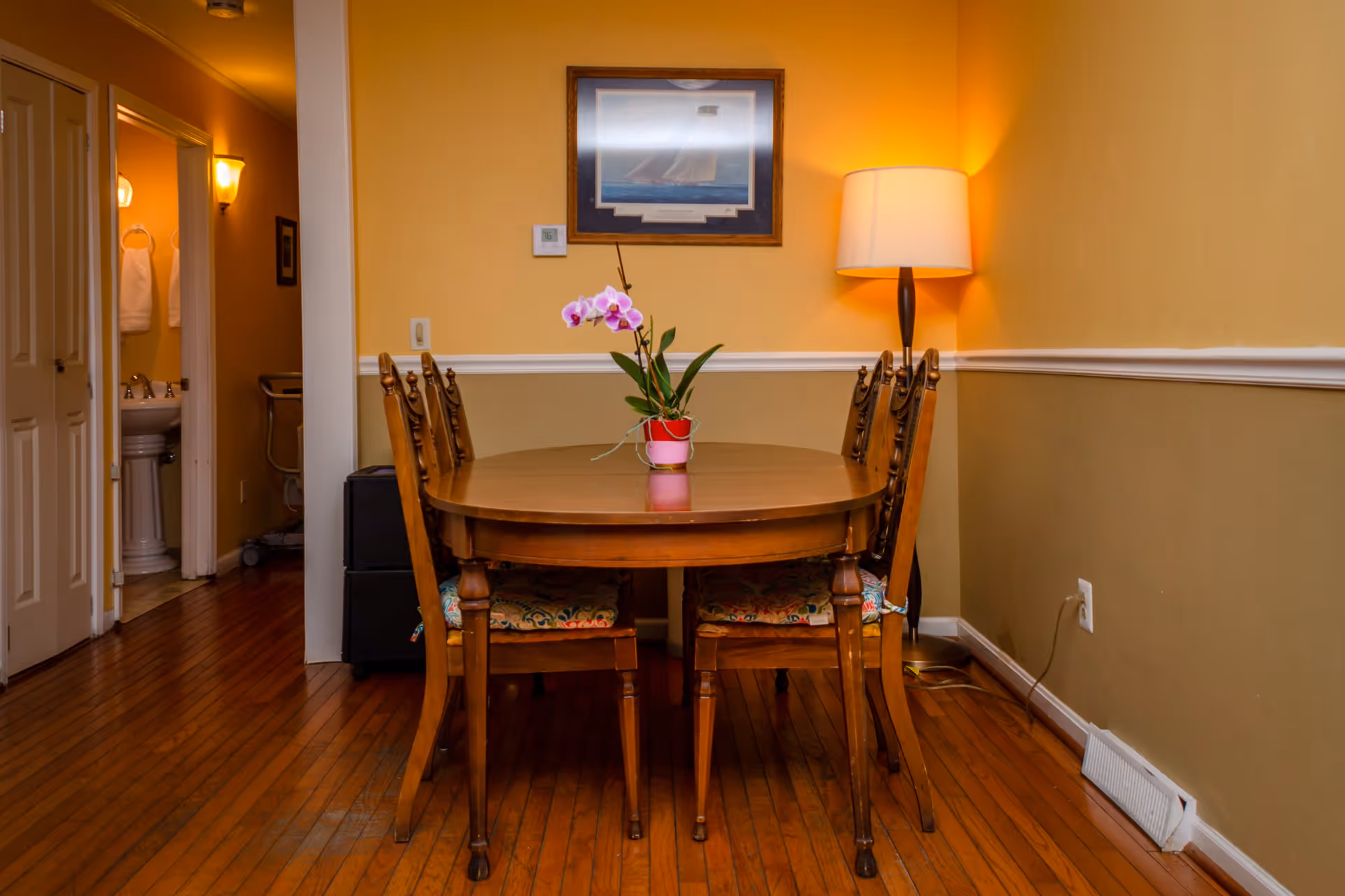 A dining room with a wooden table and six chairs, a potted orchid plant on the table, a floor lamp in the corner, and a framed picture on the yellow wall. To the left, there is an open doorway leading to a bathroom with a pedestal sink and towels hanging on the wall.