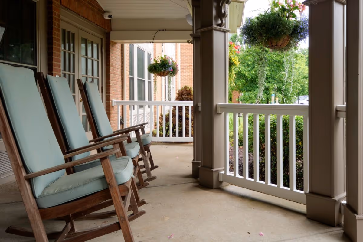 A covered porch area with three wooden rocking chairs featuring light blue cushions. The porch has white railings and hanging flower baskets with green plants and colorful flowers. The background shows a brick building and greenery outside.