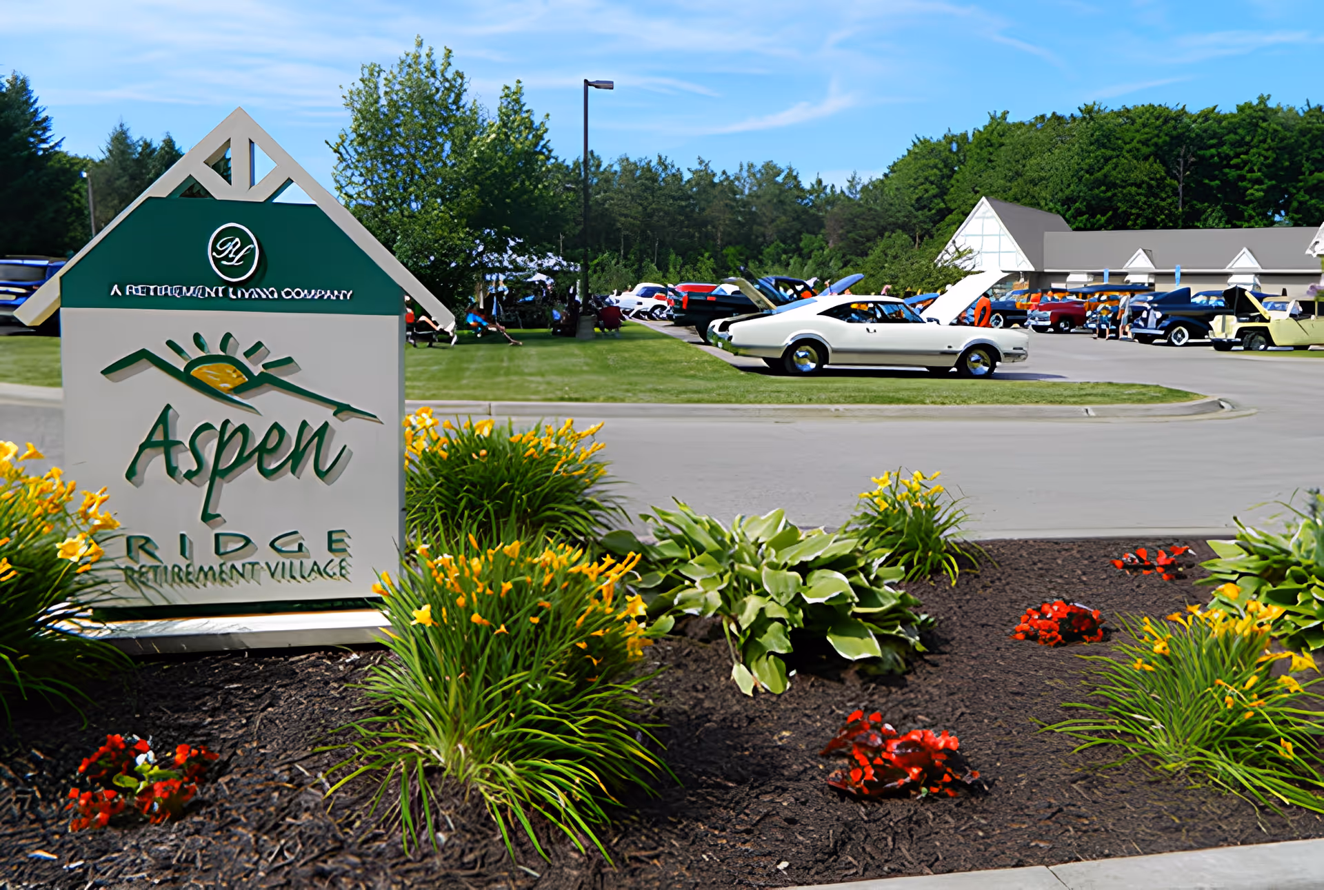 Outdoor view of Aspen Ridge Retirement Village sign surrounded by flower beds with yellow and red flowers. In the background, there is a parking area with several classic cars and a building with a white roof, set against a backdrop of green trees and a blue sky.