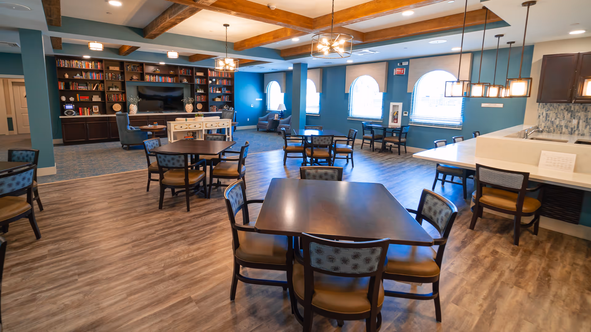 Spacious senior living common area with multiple dining tables and chairs, a kitchenette island, lounge seating and bookshelves under exposed wood-beamed ceilings.