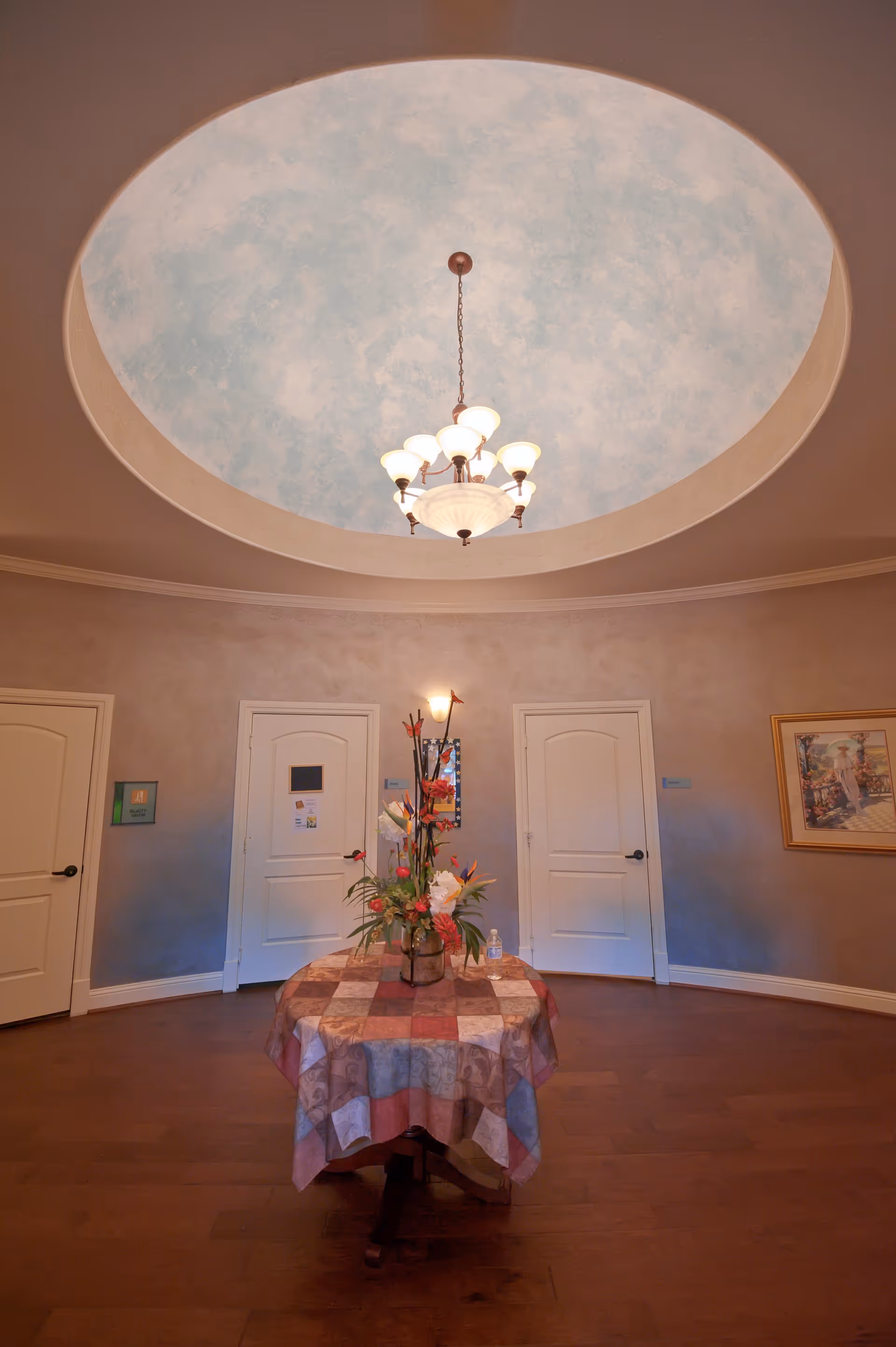 Interior view of a circular room with a round table in the center covered with a patchwork tablecloth and a floral arrangement. The room has three white doors, a painting on the wall, and a large circular recessed ceiling with a sky-like painted dome and a chandelier hanging in the middle.