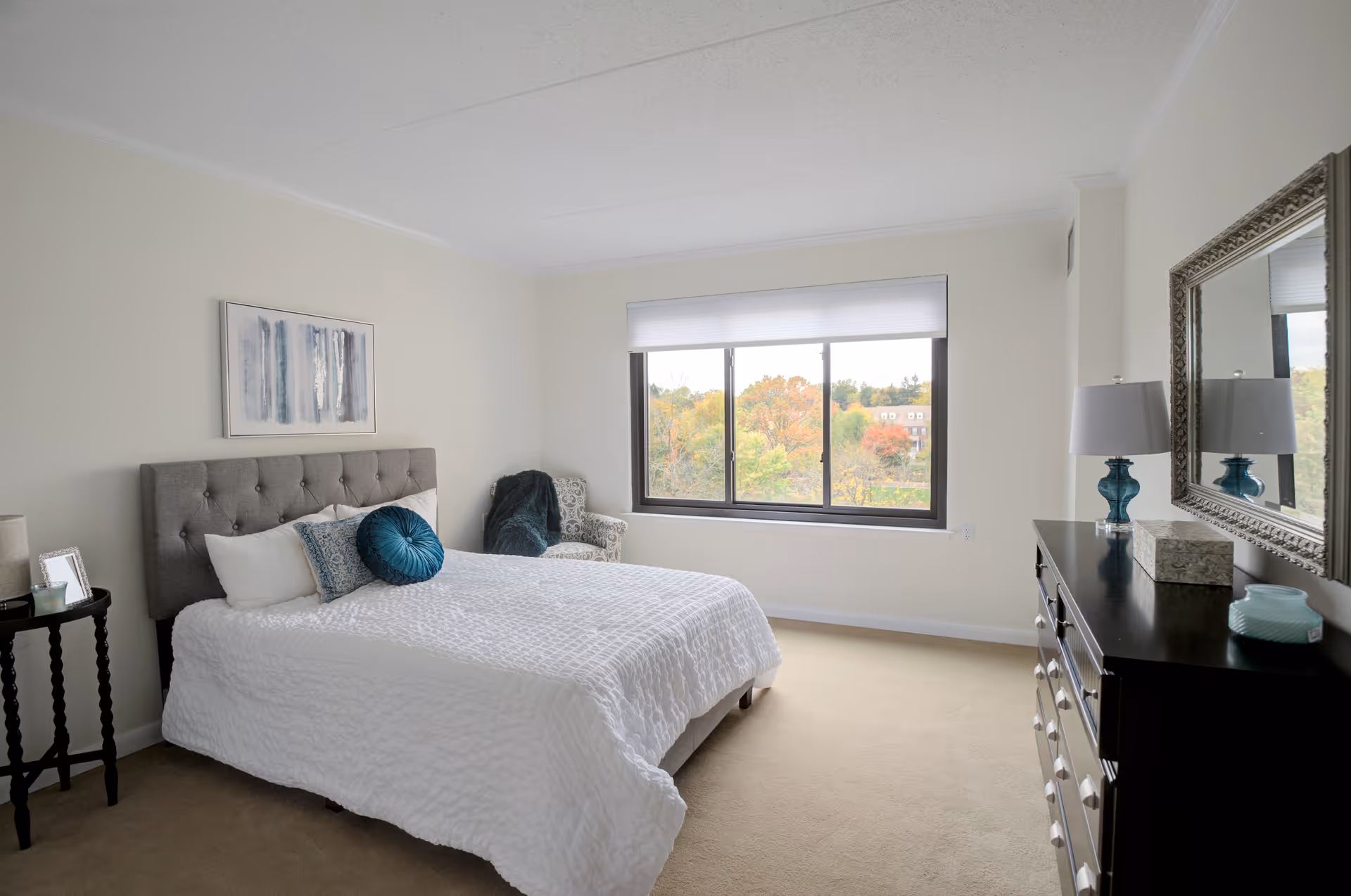 A bright and spacious bedroom with a large window showing trees outside. The room features a bed with a tufted gray headboard, white bedding, and decorative pillows. There is a small round side table with a photo frame and candle, an upholstered armchair with a throw blanket, and a dark wooden dresser with a lamp, tissue box, and decorative items. A large mirror hangs above the dresser.