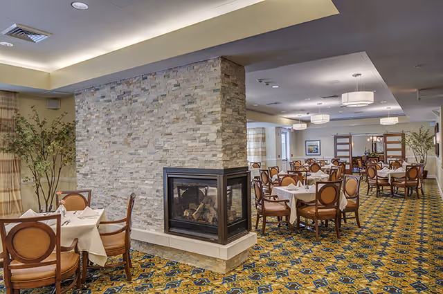 A spacious dining room in a senior living facility featuring multiple round tables covered with white tablecloths and set with napkins. The room has a patterned carpet, a stone fireplace in the center, and modern ceiling lights. There are large windows with curtains and decorative plants placed around the room.