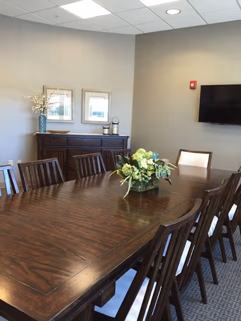A conference or dining room with a long wooden table surrounded by wooden chairs with white cushions. A floral centerpiece is placed on the table. Against the wall, there is a dark wooden sideboard with decorative items including a vase with flowers and two framed pictures above it. A flat-screen TV is mounted on the wall to the right.