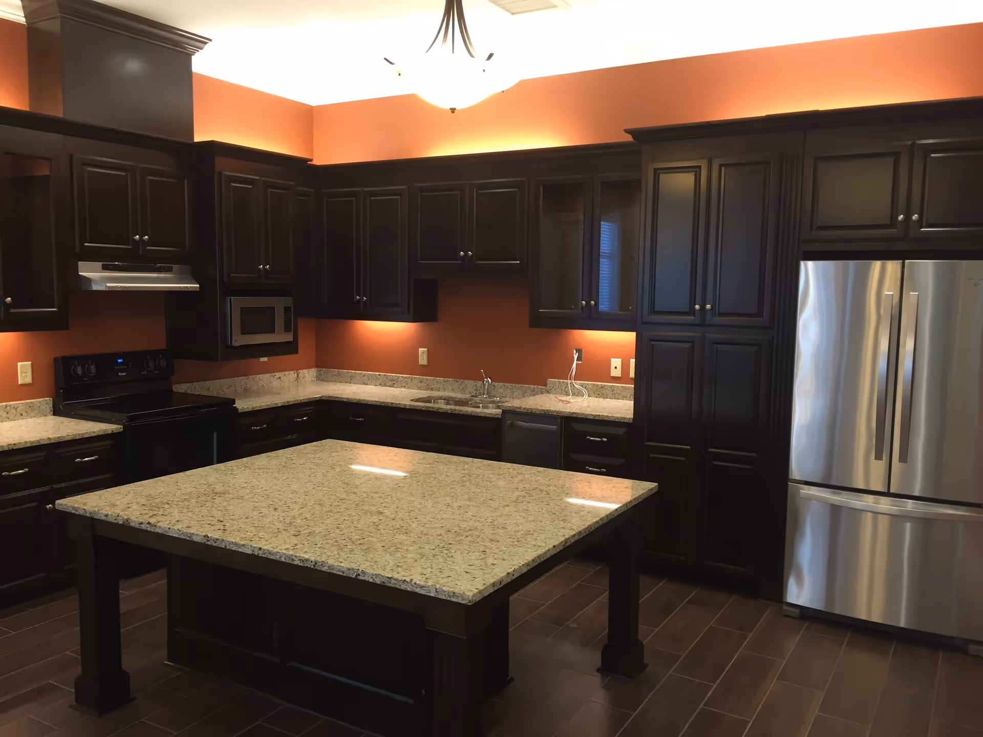 A modern kitchen with dark wood cabinets, granite countertops, a central island, stainless steel refrigerator, microwave, stove, and under-cabinet lighting. The walls are painted a warm orange color and the floor has dark brown tiles.