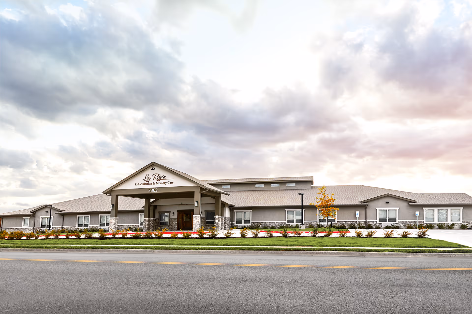 Exterior front view of Le Rêve Rehabilitation and Memory Center, a single-story building with a covered entrance, stone pillars, and landscaped greenery under a cloudy sky.