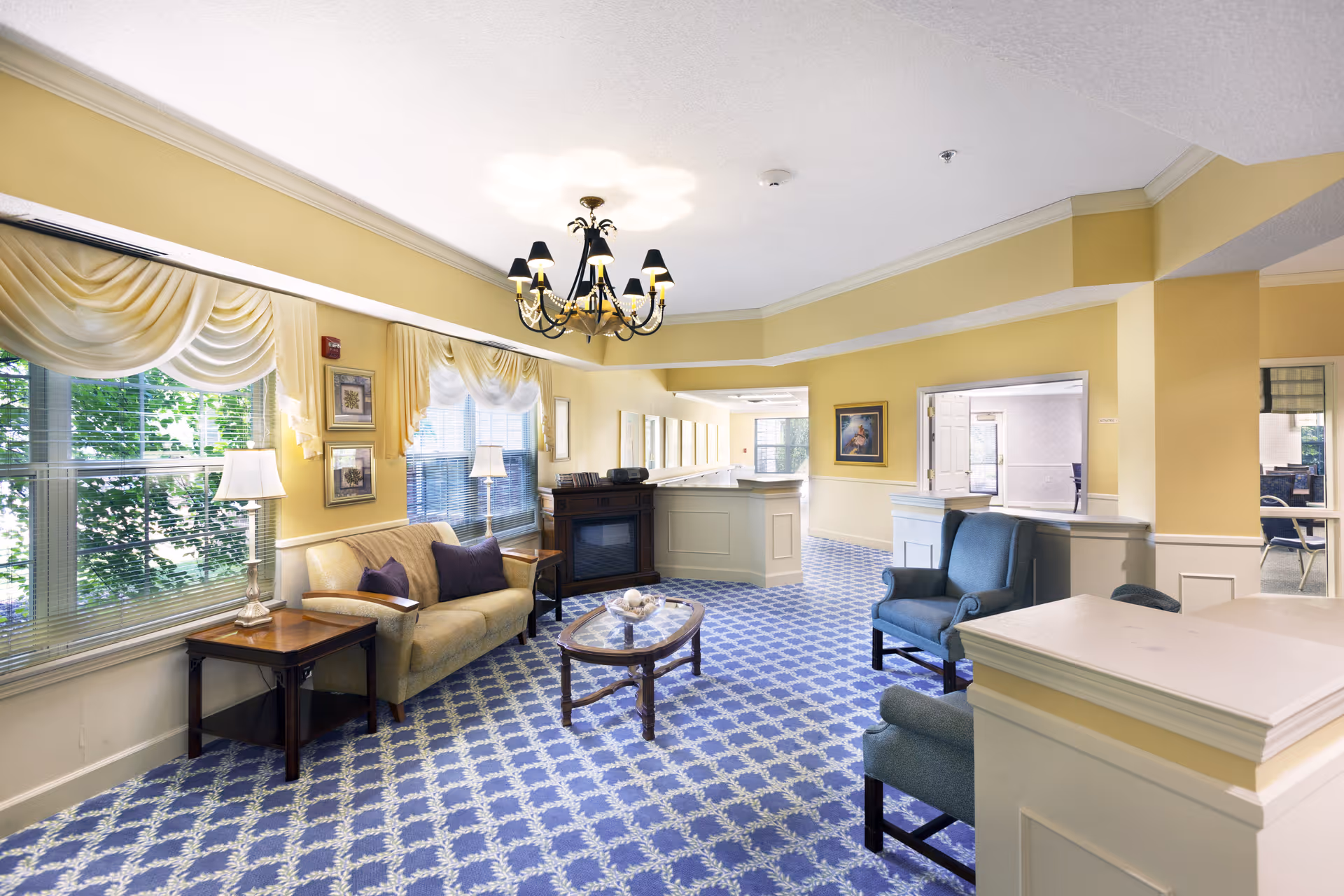 A bright and spacious living room area with yellow walls and large windows adorned with cream-colored drapes. The room features a beige sofa with dark cushions, two blue armchairs, a wooden coffee table with decorative items, and side tables with lamps. A chandelier hangs from the ceiling, and there is a fireplace against one wall. The floor is covered with a blue patterned carpet, and there are framed pictures on the walls.