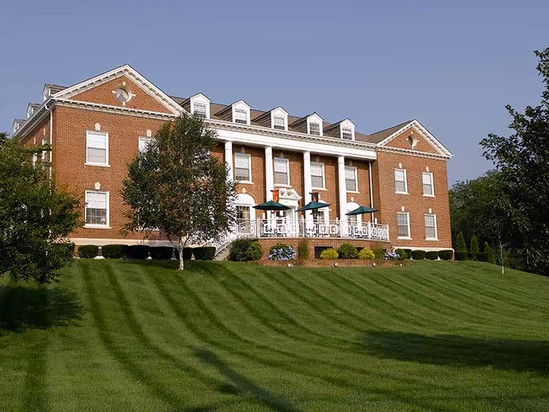 Red-brick colonial-style building with white columns, a front patio with green umbrellas, and a manicured striped lawn.