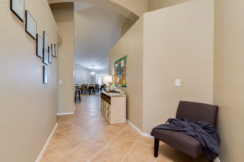 A hallway with beige walls and tiled floor leading to a dining area with a table and chairs. On the right side of the hallway, there is a small console table with baskets and a lamp on top, along with a bulletin board above it. A dark upholstered chair with a piece of clothing draped over it is positioned against the right wall. Several framed pictures or certificates are hung on the left wall.