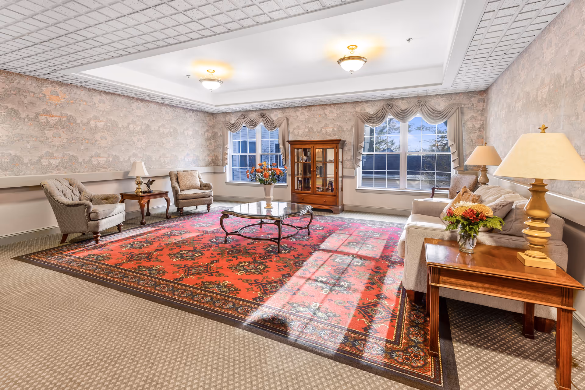 A well-lit living room with a large red patterned area rug, two armchairs, a glass-top coffee table, a wooden display cabinet, a sofa with cushions, and two side tables with lamps and flower arrangements. The room has large windows with draped curtains and wallpapered walls.