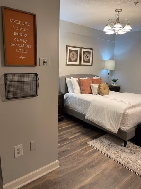 A cozy bedroom with a neatly made bed featuring white bedding and several decorative pillows in shades of white, rust, and beige. Above the bed are two framed floral artworks. A bedside table with a lamp and a small plant is visible. On the left wall, there is a wooden sign that reads 'WELCOME to our messy crazy beautiful LIFE' and a black wire basket mounted below it. The room has wood flooring and a light-colored area rug partially visible under the bed.
