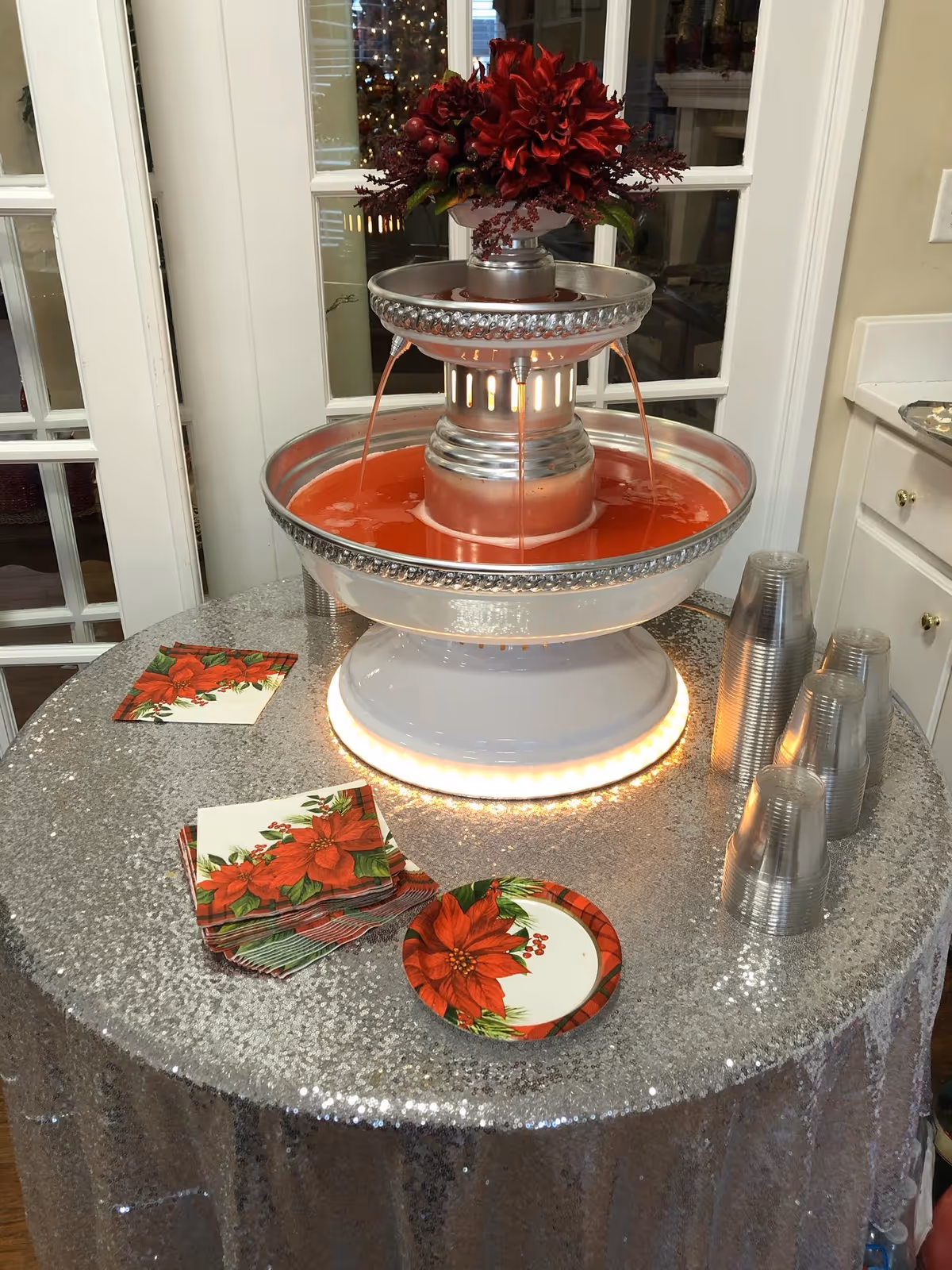 Tiered beverage fountain filled with red punch on a sequined round table with poinsettia napkins and stacked plastic cups in an interior room.