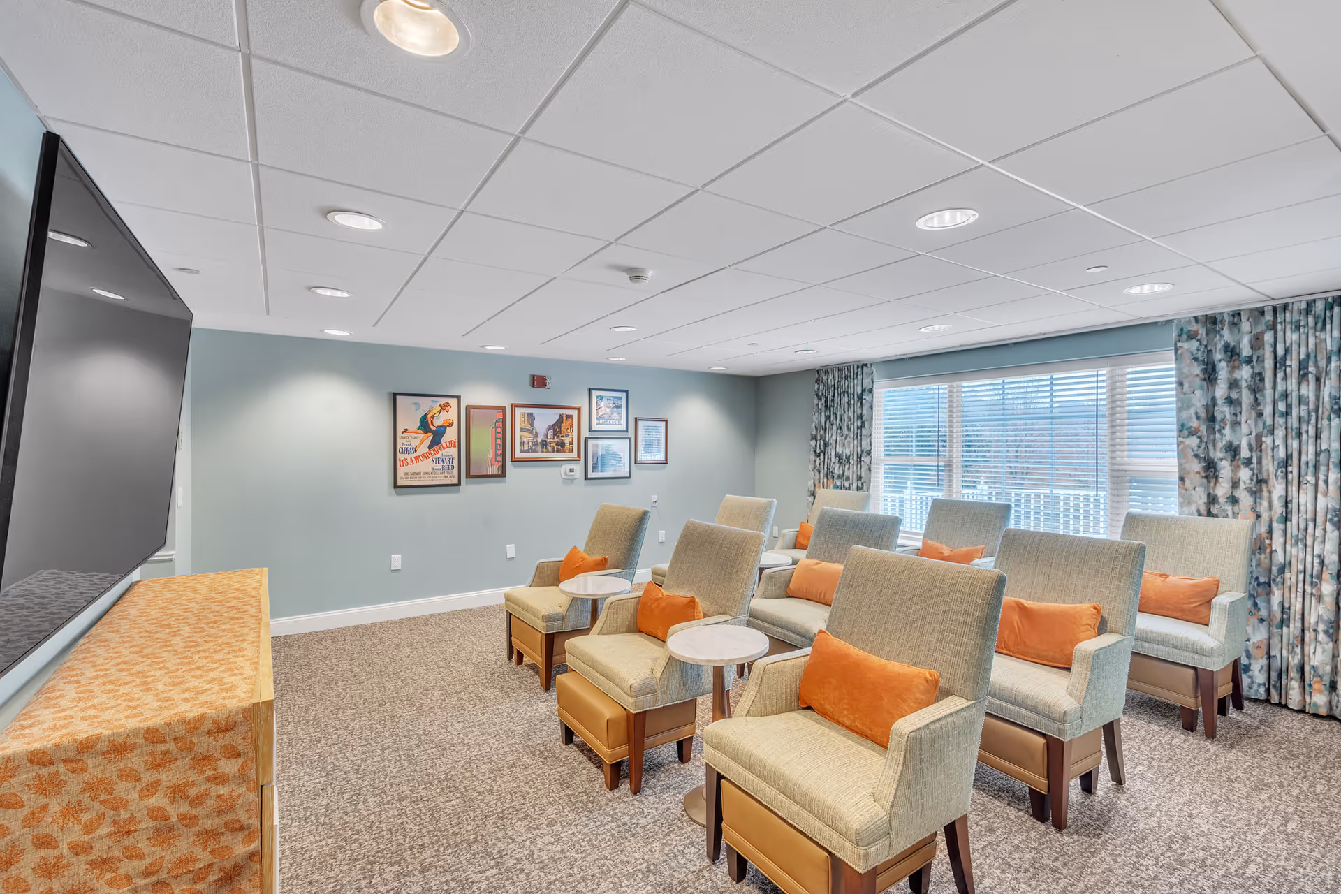 A cozy common room with multiple beige armchairs each featuring an orange cushion, arranged in rows facing a large flat-screen TV mounted on the wall. The room has light blue walls adorned with framed artwork, a large window with floral curtains letting in natural light, and a carpeted floor.