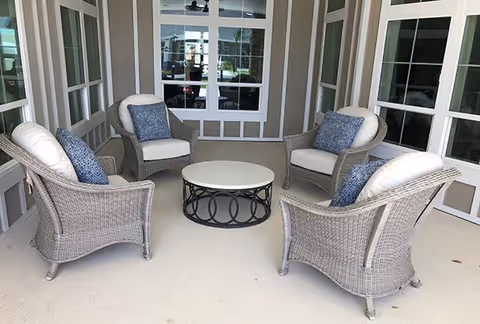 A cozy indoor seating area with four wicker armchairs arranged around a round coffee table. Each chair has white cushions and a blue patterned throw pillow. The space is surrounded by large windows and glass doors, allowing natural light to fill the room.