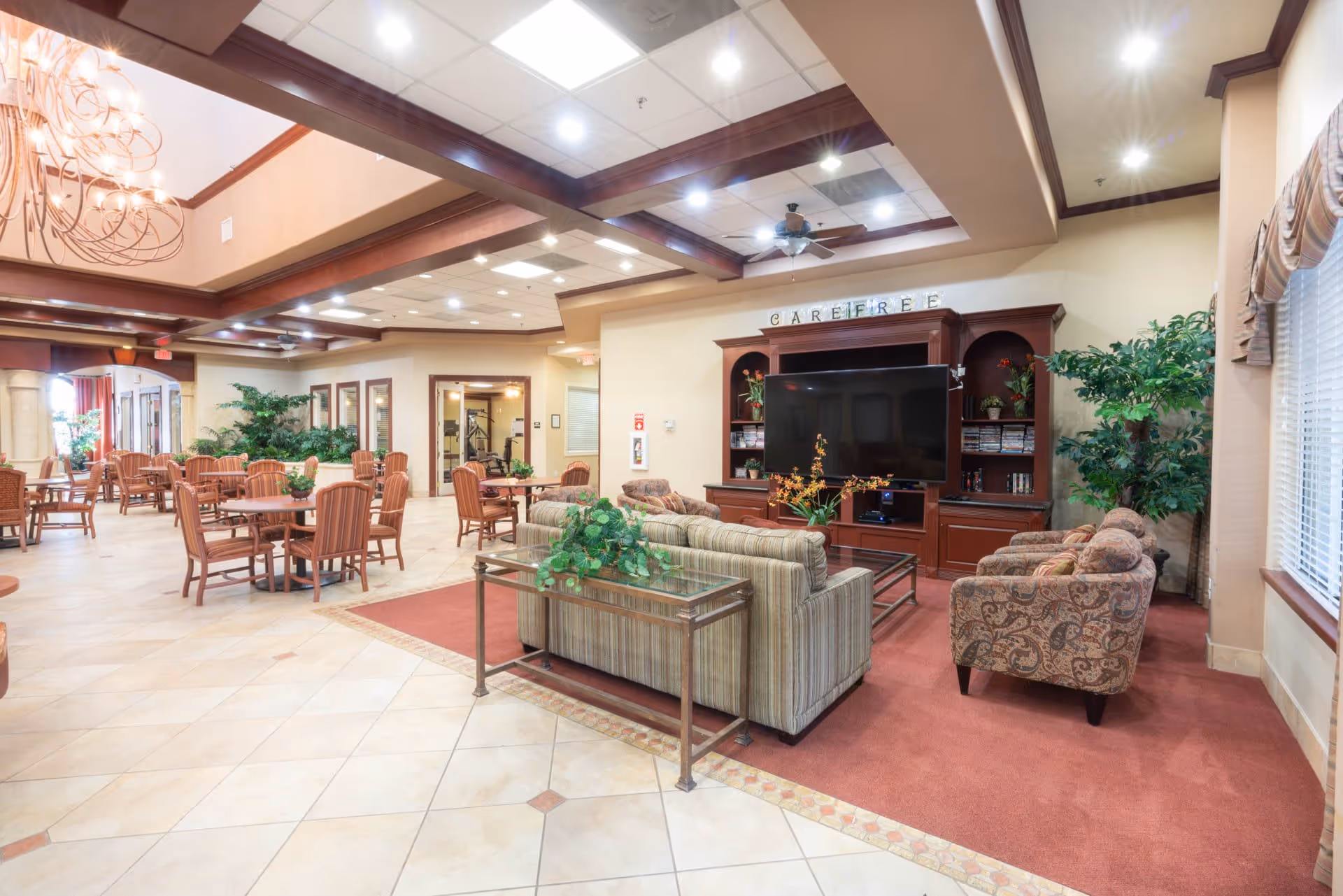 A spacious and well-lit common area in a senior living facility featuring a seating area with sofas and armchairs arranged around a large flat-screen TV on a wooden entertainment center. Several tables and chairs are set up for dining or social activities. The room has tiled floors, a high ceiling with wooden beams, and large windows with curtains allowing natural light to enter. Green plants and decorative elements add a welcoming touch.