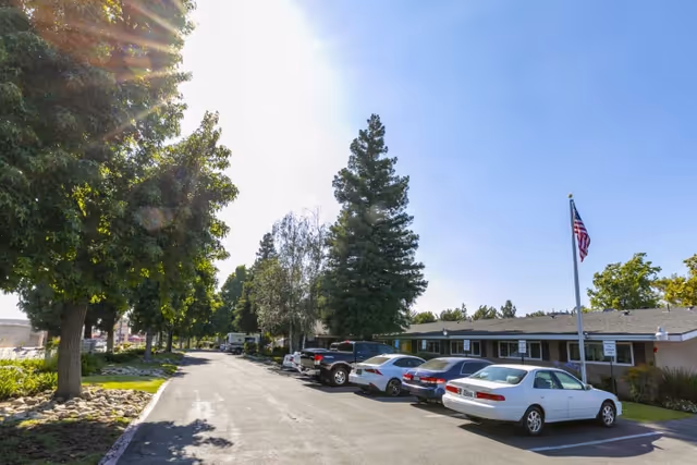 Parking lot with several cars and tall trees in front of a low single-story care facility flying an American flag.