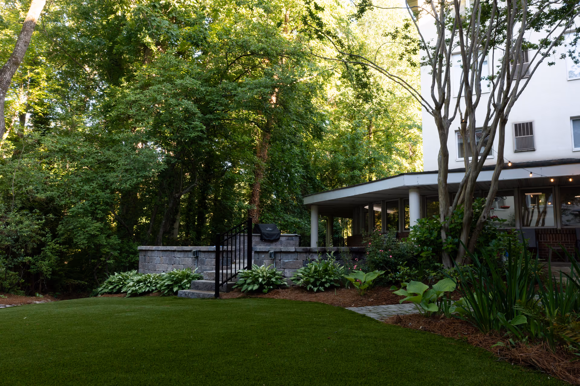 Outdoor garden area at Saint Anne's Terrace featuring a well-maintained lawn, stone steps with a black metal railing, lush green plants, and trees surrounding a covered patio attached to a white building.